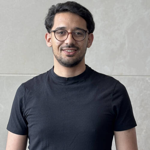 Malek Garrach, is a wearing a black  short sleeved Tshirt with a pair of spectacles on. Behind him is a gray wall